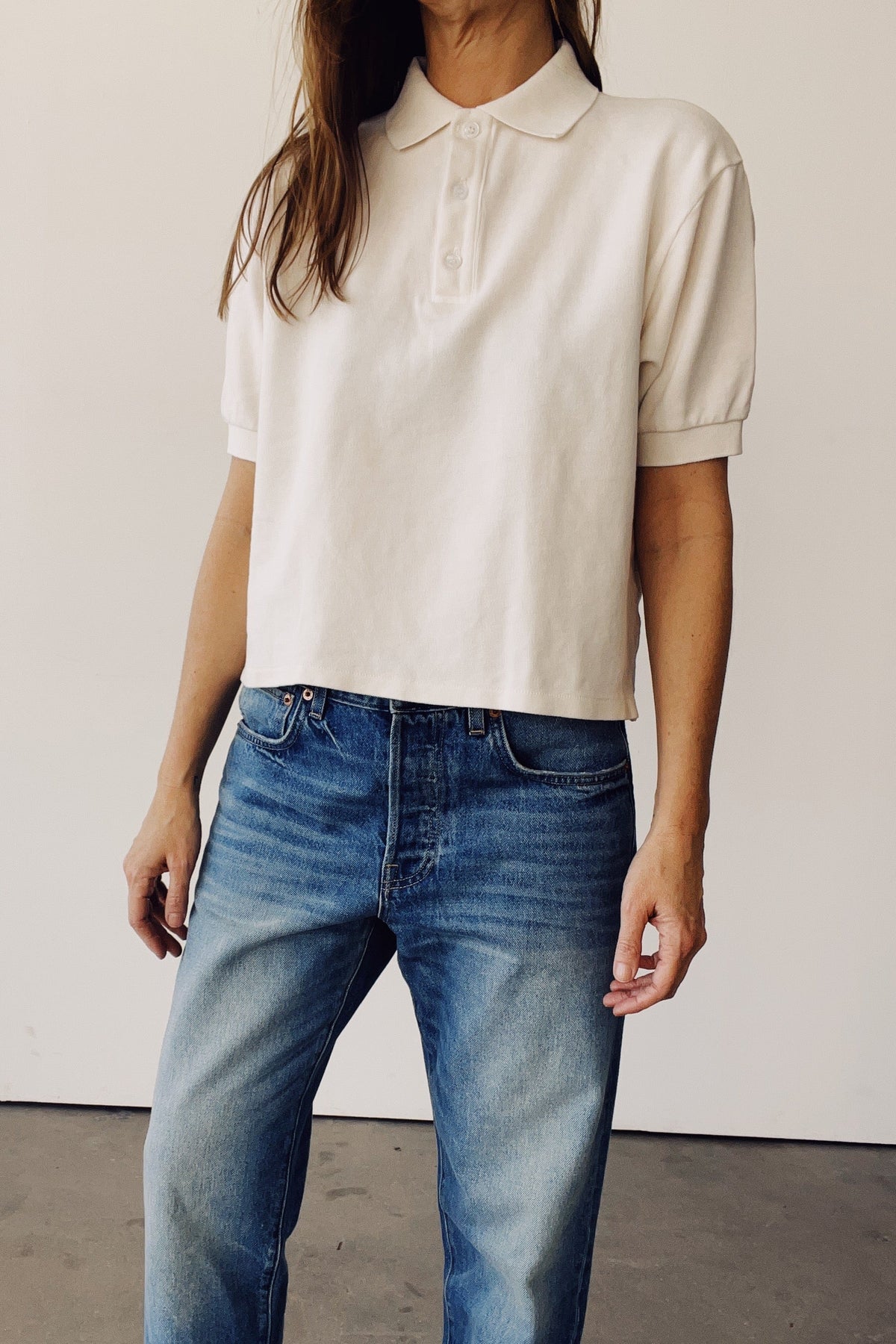 A woman wearing a short-sleeve cream-colored polo shirt and blue jeans, standing against a plain off-white wall.