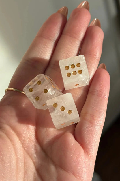 Three clear crystal dice with gold-colored dots resting on a person's palm, with a blurred indoor background.