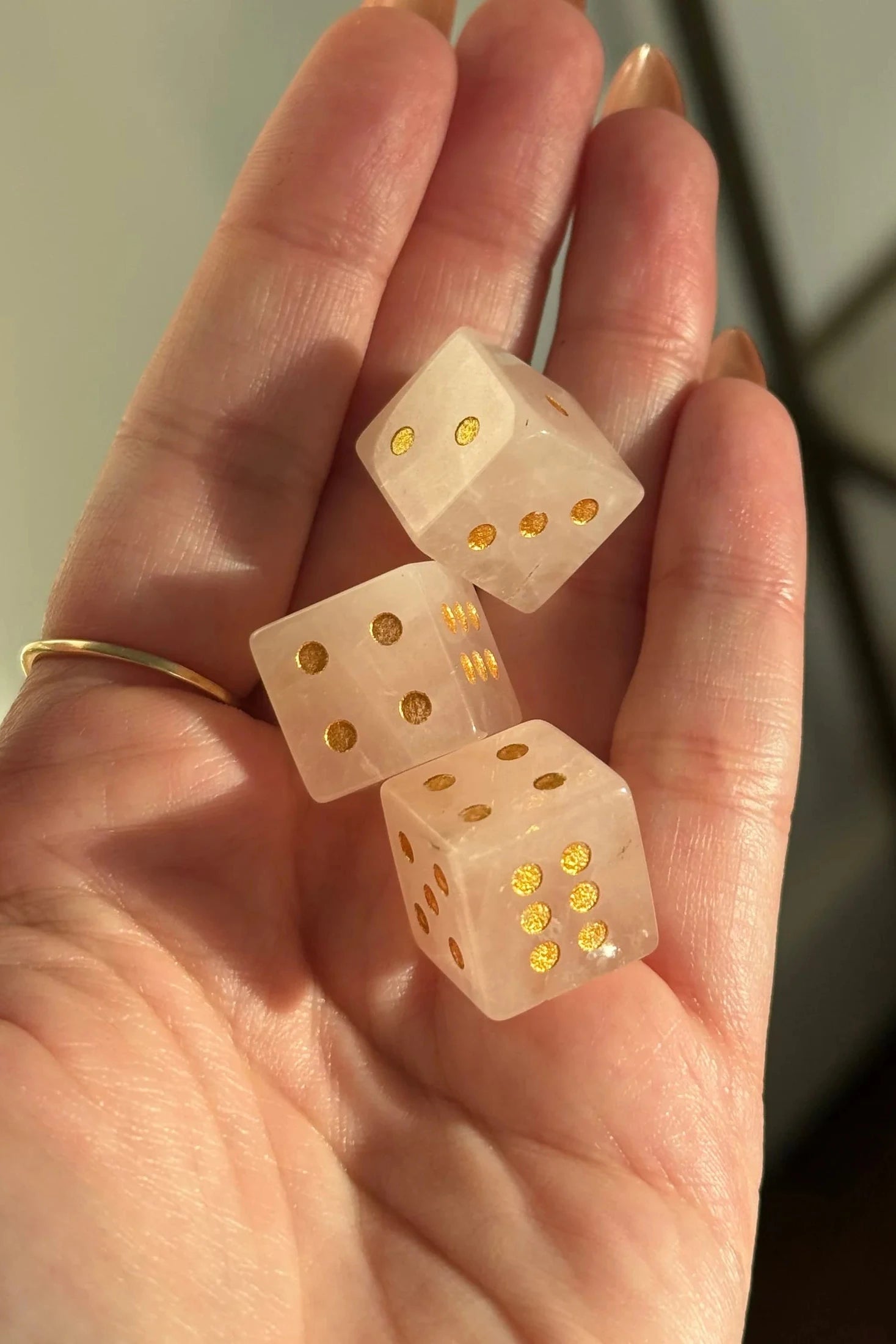 Three translucent pale pink dice with gold dot faces resting on an open hand. The hand has a gold ring on one finger, and the background is a softly blurred indoor setting.