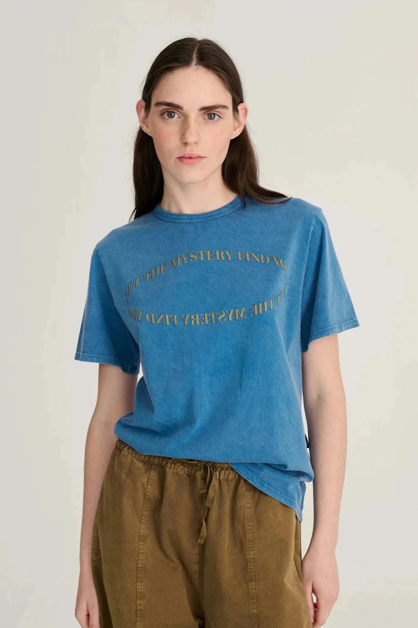 A woman modeling a blue short-sleeve graphic t-shirt with gold text, paired with brown pants, against a plain light gray background.
