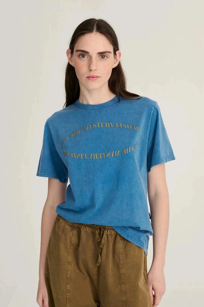 A woman modeling a blue short-sleeve graphic t-shirt with gold text, paired with brown pants, against a plain light gray background.