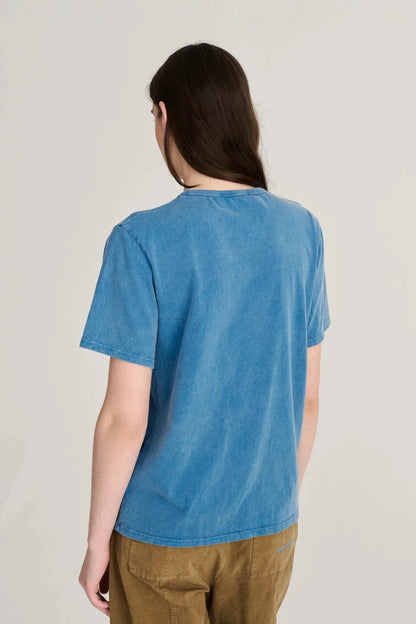 A model shown from the back wearing a short-sleeve blue t-shirt and brown pants against a plain light gray background.