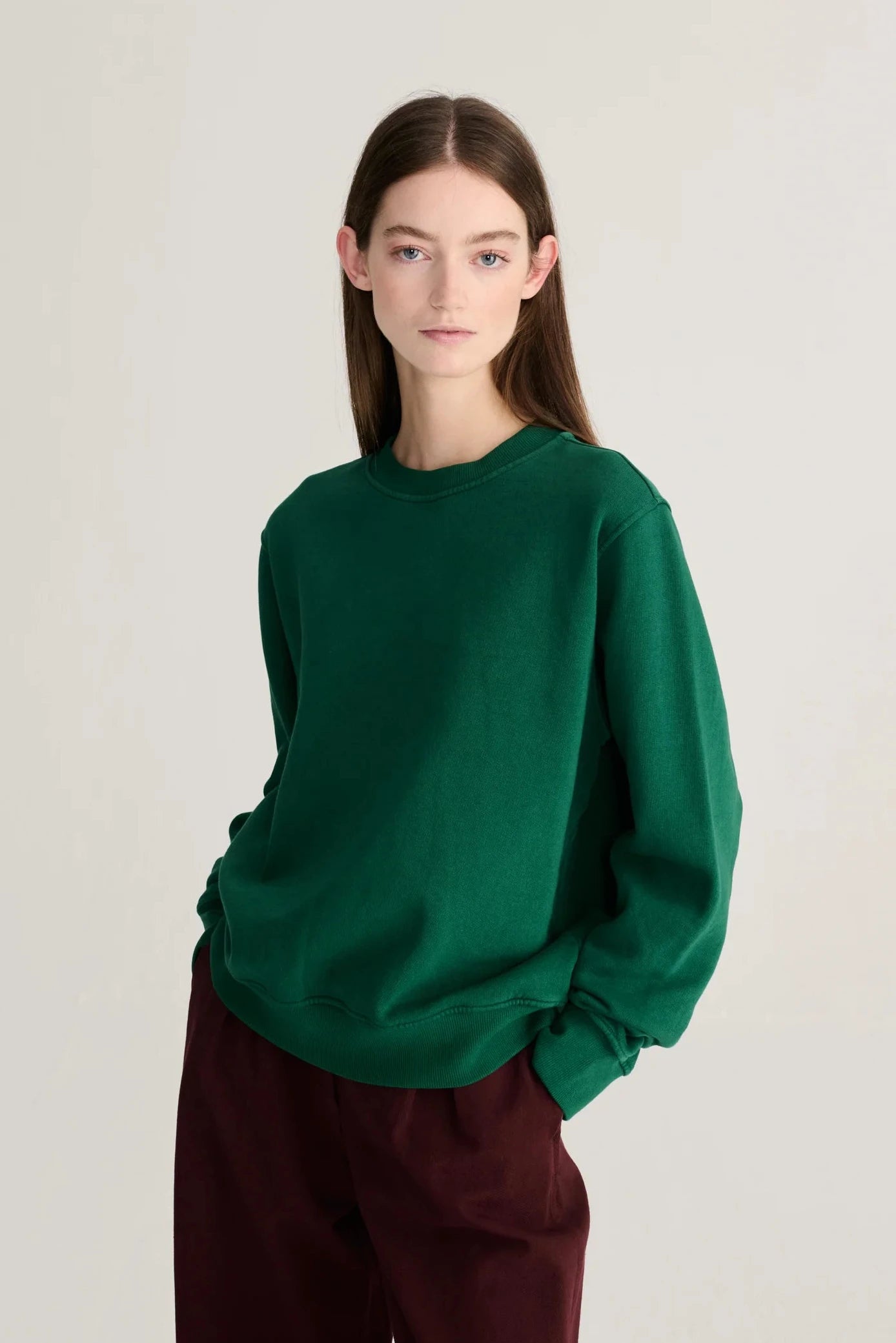 A model wearing a dark green crewneck sweatshirt and dark burgundy pants, standing against a plain light gray studio background.