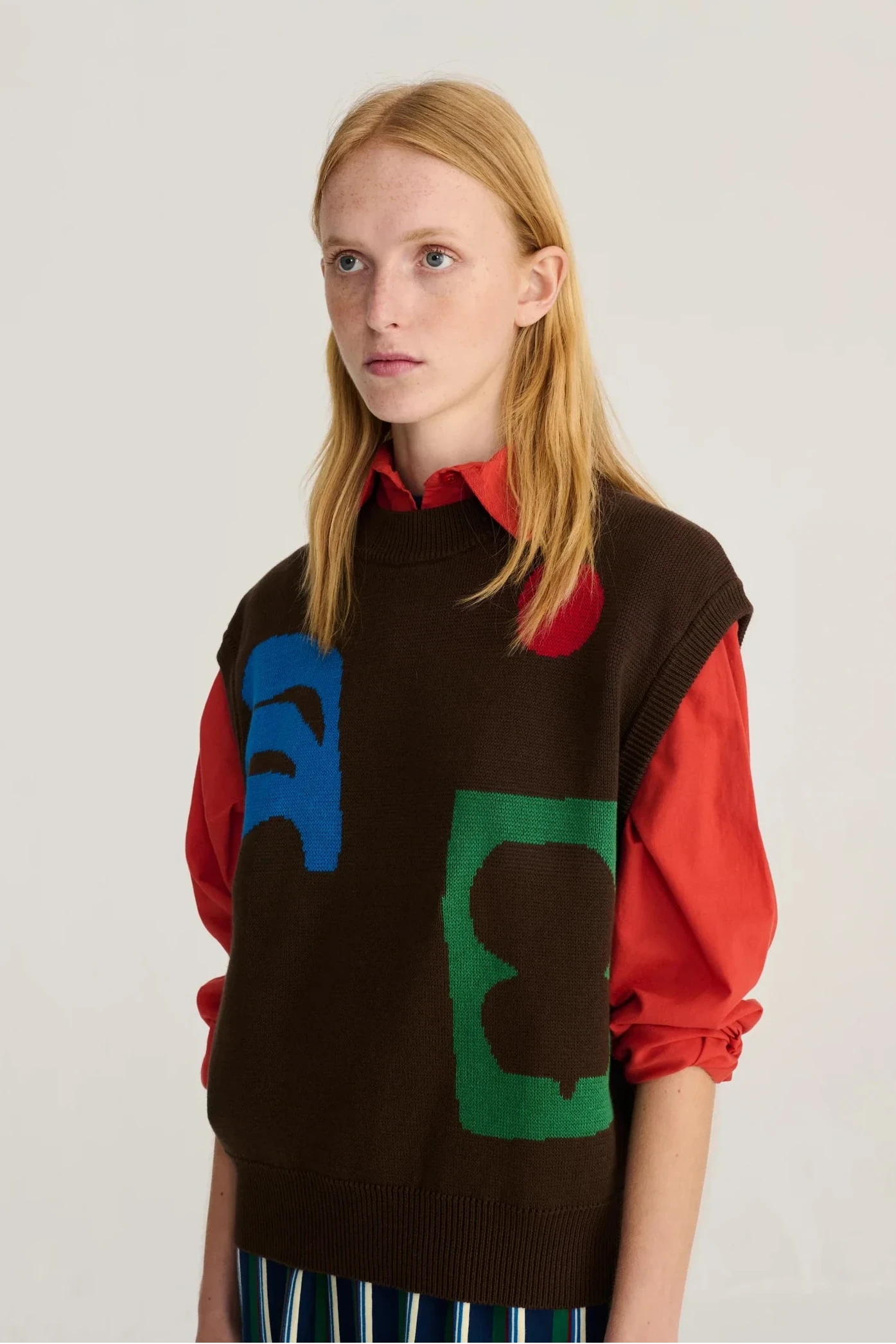 A blonde female model wearing a brown mock-neck knit sweater vest with red sleeves and large abstract blue, green, and red graphic shapes on the front, paired with a red collared shirt and striped trousers, against a plain light gray background.