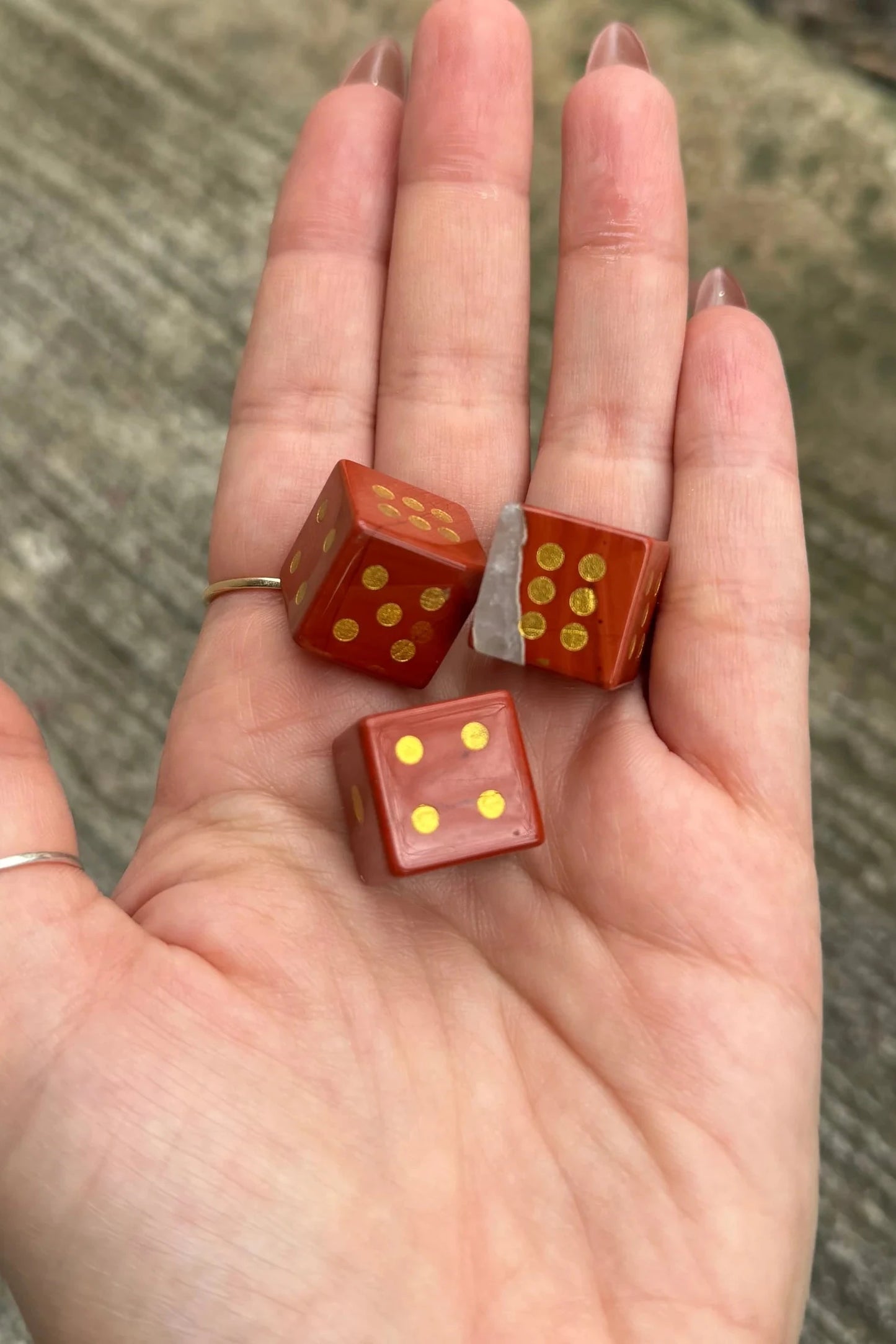 Three small red dice with gold dots resting on an open hand, photographed outdoors against a blurred gray concrete or stone surface.