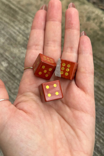 Three small red dice with gold dots resting on an open hand, photographed outdoors against a blurred gray concrete or stone surface.
