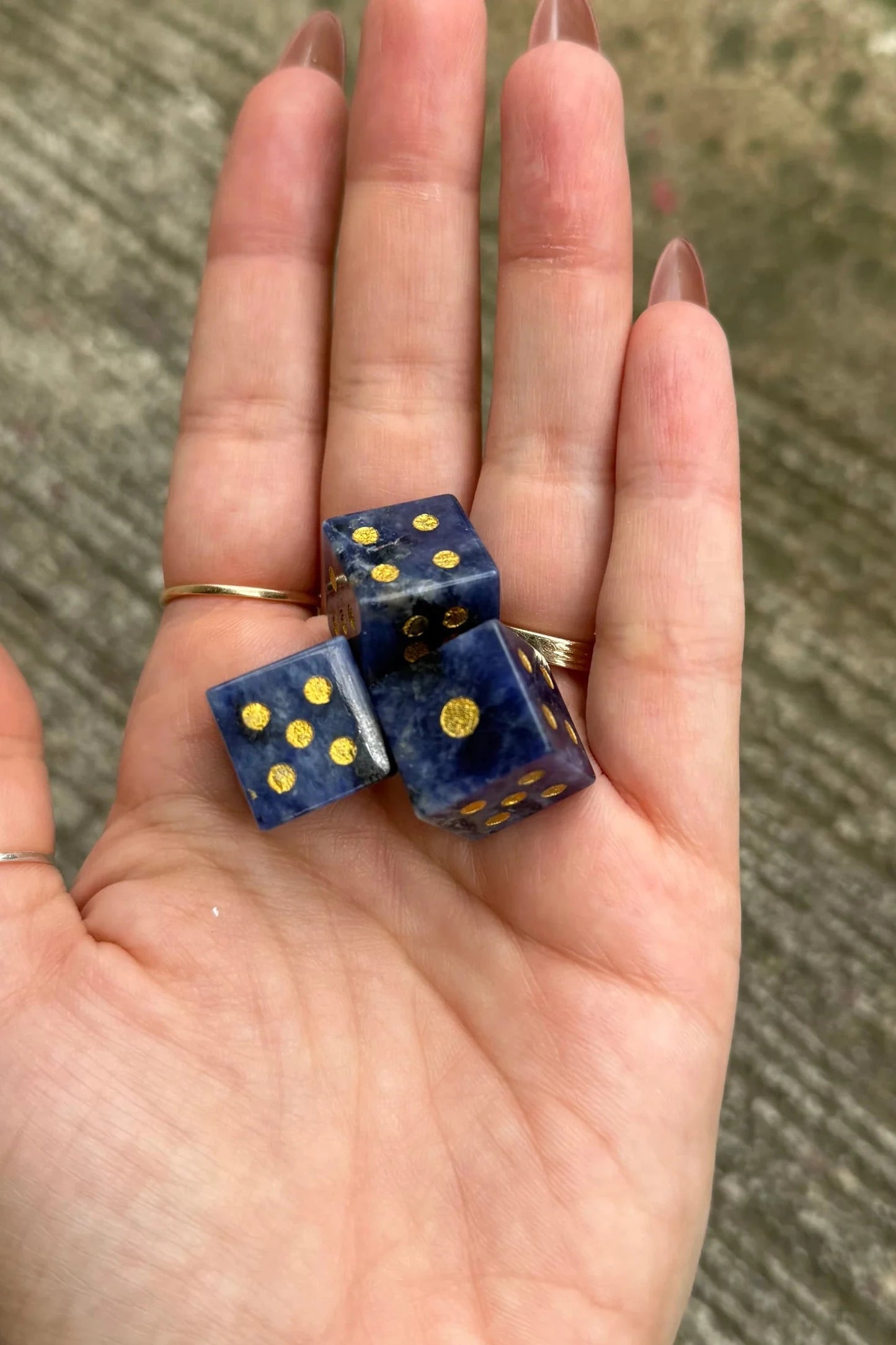 Three blue and gold gemstone dice resting on an open hand, with a blurred concrete surface in the background.
