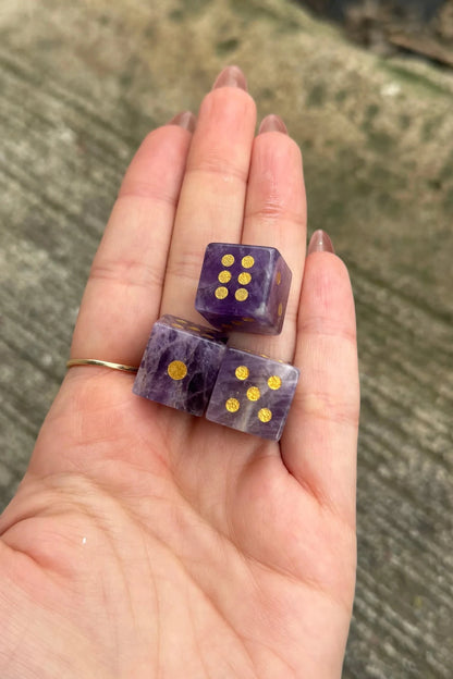 Three purple amethyst dice with gold foil dot markings resting on an open hand, with a blurred outdoor concrete background.