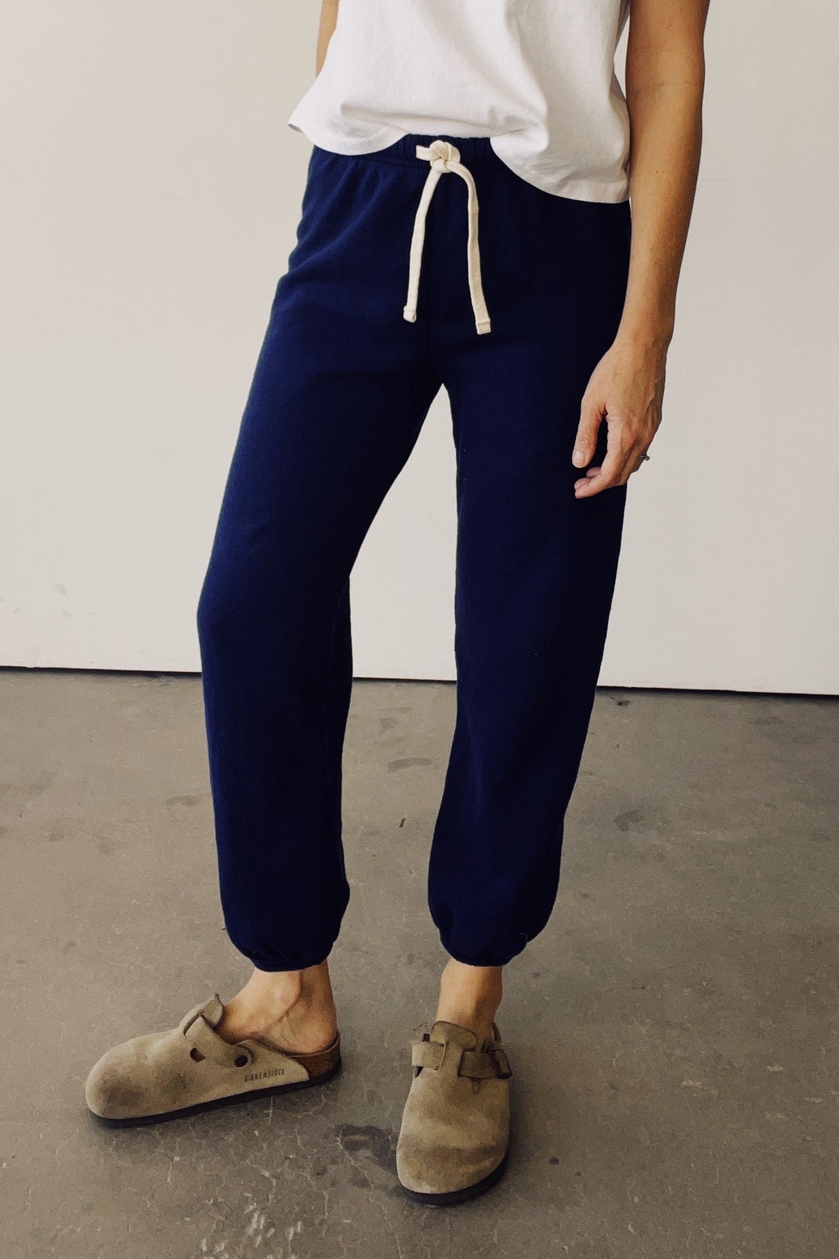 A person wearing navy jogger-style pants with a cream drawstring, paired with a white top and taupe slip-on clogs, standing against a plain off-white wall and concrete floor.