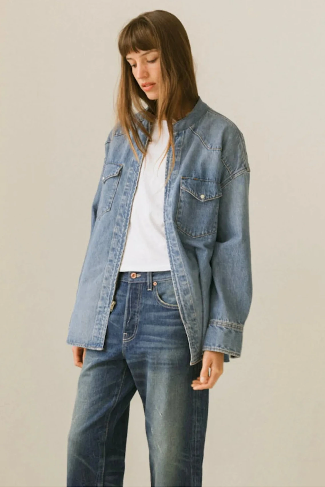 A woman wearing a light-wash blue denim jacket over a white t-shirt and dark blue jeans, standing against a plain light beige studio background.