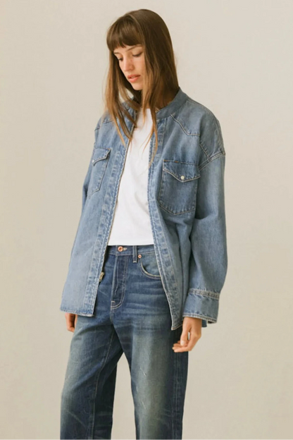 A woman wearing a light-wash blue denim jacket over a white t-shirt and dark blue jeans, standing against a plain light beige studio background.