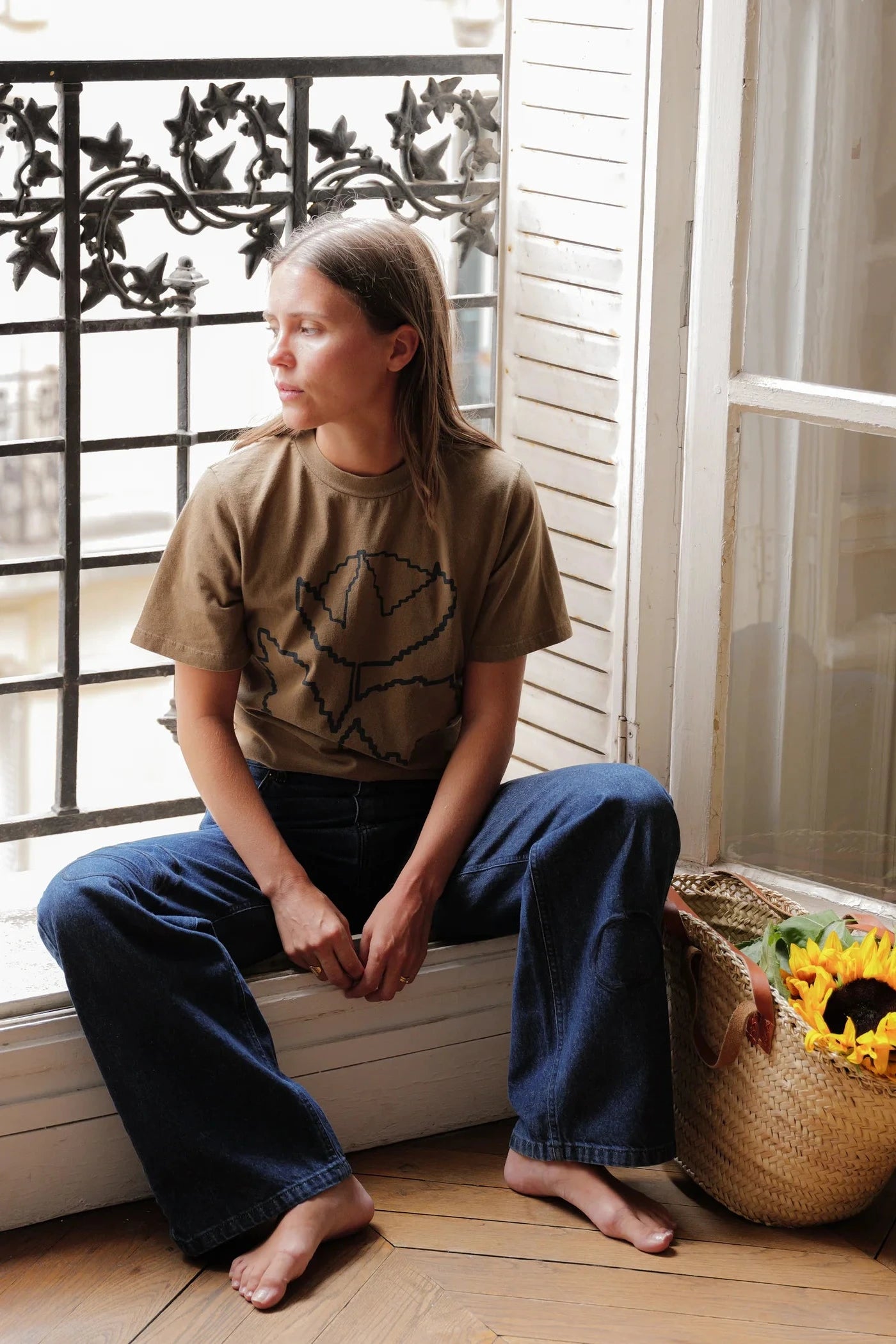A woman sits barefoot on a windowsill in a sunlit room, wearing a brown graphic T-shirt and loose dark blue jeans. A woven basket of sunflowers rests beside her near the window.