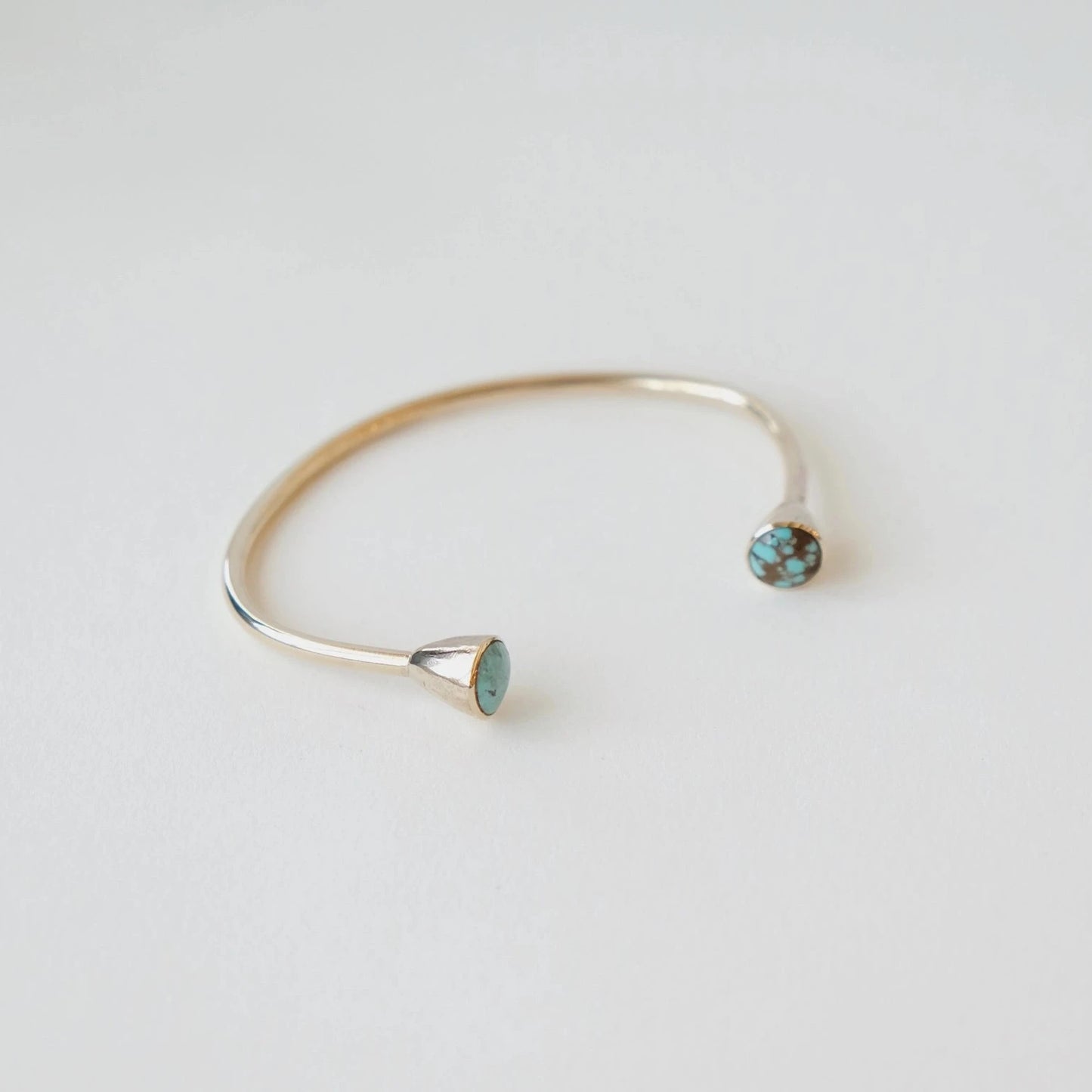 A minimalist open cuff bracelet in polished gold-tone metal, featuring two small bezel-set round turquoise-blue gemstones, photographed on a clean white background.